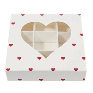 Puede incluir: Caja de cartón blanca con una ventana en forma de corazón y un patrón de corazones rojos. La caja tiene nueve compartimentos en el interior.
