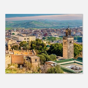 May include: An aerial view of a city in Morocco with a tall, brown tower in the foreground. The tower has a pointed roof and is surrounded by trees and buildings. In the background, the city stretches out across a valley with mountains in the distance.