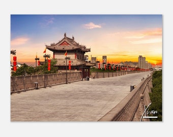 May include: A scenic photograph of a historic Chinese city wall and gatehouse under a vibrant sunset. The image features a long, gray stone pathway, red lanterns, and traditional architecture. The sky transitions from blue to orange.