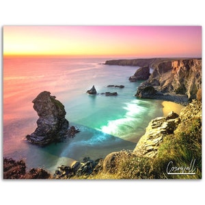 May include: A panoramic view of a rocky coastline with a sandy beach and turquoise water. The sky is a vibrant pink and orange, creating a beautiful sunset scene. The image is captioned "Cornwall" in the bottom right corner.