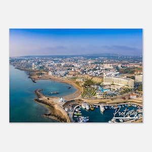 Puede incluir: Una vista aérea de una ciudad costera con un puerto lleno de barcos. La ciudad está construida en una península con una pequeña playa y un mar azul claro. El texto "Protaras" está escrito en blanco sobre un fondo azul en la esquina inferior derecha de la imagen.
