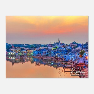 May include: A panoramic view of a city skyline at sunset, with a lake in the foreground. The city is built on a hill and features many buildings with blue and white facades. The sky is a vibrant orange and pink, reflecting in the water.