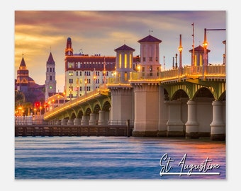 May include: A view of the Bridge of Lions in St. Augustine, Florida at sunset. The bridge is lit up with golden lights and the city skyline is visible in the background.