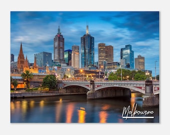 Könnte beinhalten: Ein Panoramablick auf die Skyline von Melbourne in der Abenddämmerung, mit einer Brücke über einen Fluss und den Wolkenkratzern der Stadt im Hintergrund. Die Lichter der Stadt spiegeln sich im Wasser und schaffen eine wunderschöne und lebendige Szene. Der Text "Melbourne" ist in weißer Kursivschrift am unteren Rand des Bildes geschrieben.
