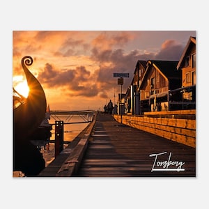 May include: A wooden dock extends into the water at sunset, with a silhouette of a Viking ship in the foreground. The sky is a vibrant orange and pink, and the water is calm and reflective. The dock is lined with wooden buildings, and the sign "Soppel" is visible in the distance. The text "Tonsberg" is written in white on the dock.