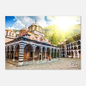 Könnte beinhalten: Ein farbenfrohes, historisches Kloster mit schwarz-weißen gestreiften Bögen, eingebettet in eine Berglandschaft. Die Sonne scheint hell am Himmel und beleuchtet das Gebäude. Der Text "Rila" ist in weiß auf schwarzem Hintergrund in der unteren rechten Ecke des Bildes geschrieben.