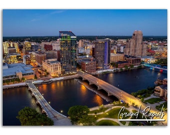 May include: An aerial view of Grand Rapids, Michigan at dusk. The city skyline is illuminated with lights, and the Grand River flows through the center of the image. Several bridges cross the river, and there are trees and green spaces along the riverbanks. The text "Grand Rapids" is written in cursive in the lower right corner of the image.