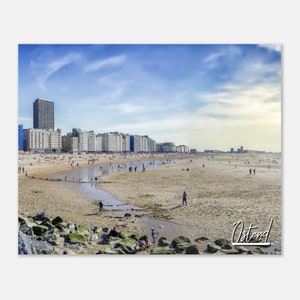 Może przedstawiać: Panoramiczny widok plaży w Ostendzie w Belgii. Plaża jest głównie piaszczysta, z kilkoma osobami spacerującymi i bawiącymi się w wodzie. W oddali widać budynki, a niebo jest błękitne z białymi chmurami.