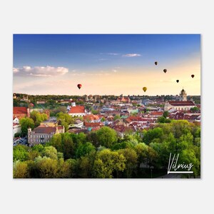 Puede incluir: Una vista aérea de la ciudad de Vilnius, Lituania, con globos aerostáticos flotando en el cielo. La ciudad es una mezcla de edificios antiguos y modernos, con muchos árboles verdes y vegetación. El cielo es de un azul hermoso con algunas nubes. El texto "Vilnius" está escrito en blanco en la esquina inferior derecha de la imagen.