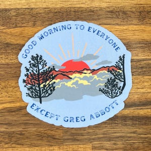 May include: A light blue sticker with a sunrise design and the text "GOOD MORNING TO EVERYONE EXCEPT GREG ABBOTT" in blue. The design features a red sun, orange rays, and silhouettes of trees.