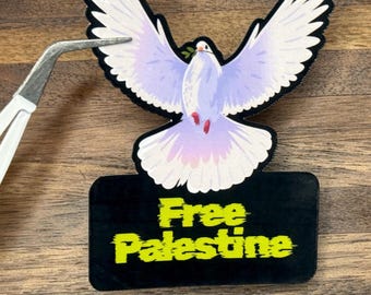 May include: A sticker featuring a dove with outstretched wings, holding an olive branch. The dove is light purple with white and pink accents. Below the dove is a black rectangle with the words "Free Palestine" in yellow.