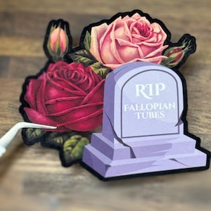 May include: A sticker featuring a purple tombstone with the text "RIP FALLOPIAN TUBES" and a bouquet of roses in shades of pink and red. The sticker has a black border and is placed on a wooden surface.