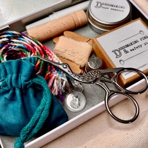 May include: A sewing kit containing a pair of silver stork-shaped scissors, a teal drawstring pouch, a spool of multicoloured braided thread, a wooden tool, a metal thimble, a round tin labelled Dressmaking tape measure, and a box labelled Dressmaking Pins.