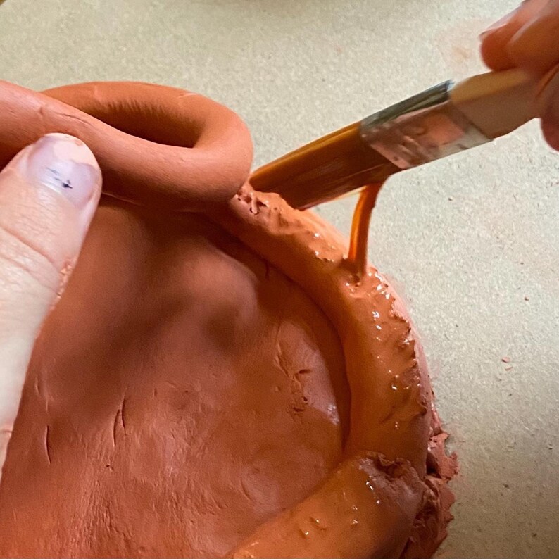 May include: A close-up of a terracotta clay pot being glazed with a brush. The pot is a warm orange color, and the glaze is a glossy, wet orange. A hand is holding the brush, applying the glaze to the rim of the pot.