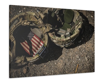 May include: An overhead shot of two military helmets with camouflage patterns. One helmet contains a folded American flag. The helmets are resting on a gravelly surface, with a small, thin, wooden stick visible.