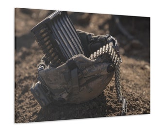 May include: A canvas print featuring a close-up of military equipment. The image shows a camouflage-patterned ammunition carrier filled with bullets, with a belt of bullets extending from it. The scene is set on a dirt ground.