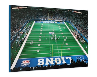 May include: A canvas print of a football field with the words "LIONS" and "NFL" visible. The field is green with white yard lines and numbers. The stands are filled with spectators. The canvas has a blue border.