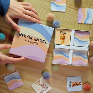Op de afbeelding: Een memory-spelset met de tekst "TIERISCHE GEFÜHLE MEMO-SPIEL" op de doos. De doos en kaarten hebben pastelkleurige golfontwerpen en dierenillustraties. Kleurrijke pompons zijn verspreid. Het spel staat op een houten oppervlak.