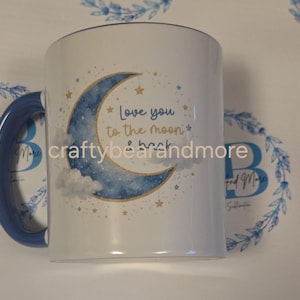 Pode incluir: Uma caneca de cerâmica branca com alça azul, apresentando uma ilustração em aquarela de uma lua crescente, nuvens e estrelas. O texto "Love you to the moon & back" está impresso em azul e dourado no centro do design da lua.