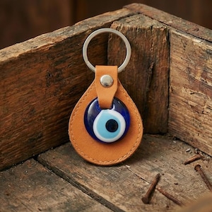 May include: A tan leather keychain with a blue and white evil eye charm. The keychain has a silver metal ring and is set against a rustic wooden background. The charm is a teardrop shape.