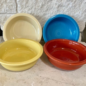 May include: Four colorful ceramic bowls in cream, turquoise, yellow, and red. The bowls are round with a slightly flared rim and a concentric circle design in the center. The bowls are arranged on a concrete surface.