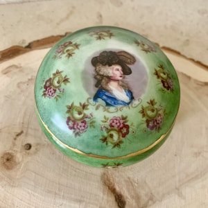 May include: A round, green porcelain trinket box with a gold trim. The lid features a painted portrait of a woman in a blue dress and a black hat, surrounded by floral designs.