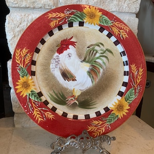 May include: A red and white ceramic plate with a painted image of a rooster, sunflowers, and wheat. The plate is on a metal stand.