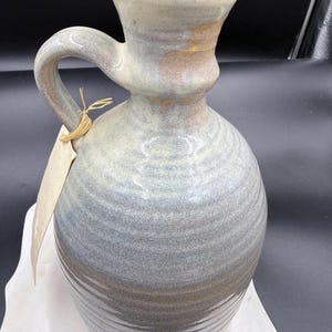 May include: A handmade ceramic pitcher with a cork stopper. The pitcher has a rounded body and a curved handle. The colors are a blend of gray, blue, and brown. A blank tag is tied to the handle with twine. The pitcher is sitting on a white surface.