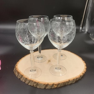 May include: Set of four clear glass wine glasses with etched floral designs. The glasses are arranged on a wooden round slice.