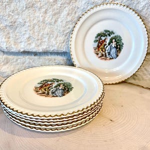 May include: A set of six white ceramic plates with a gold trim and a delicate floral design featuring a couple in a garden setting.