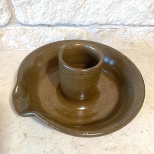 May include: A brown ceramic dish with a smaller brown ceramic cup in the center. The dish has a lip on one side and a slightly raised edge around the cup.