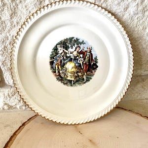 May include: A white ceramic plate with a gold rim and a vintage illustration of a couple dancing in a garden setting.