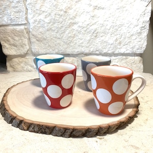 May include: Four ceramic mugs with white polka dots on a wooden serving tray. The mugs are red, orange, teal, and gray. The tray is a natural wood slice with a rough bark edge. The mugs have white interiors and white handles.