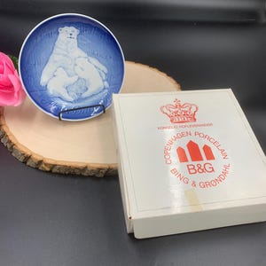 May include: A blue and white porcelain plate with a depiction of a polar bear and two cubs. The plate is in its original box, which features the Royal Danish Crown and the text "KONGELIG HOFLEVERANDØR", "COPENHAGEN PORCELAIN", "BING & GRØNDAHL", and "B&G".