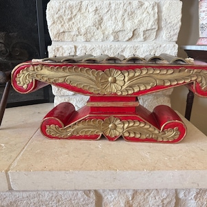 May include: A red and gold gamelan instrument with a decorative floral design. The instrument has a series of metal bars and a wooden mallet. The base is red with gold accents.