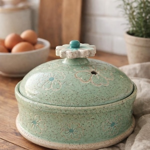 May include: A ceramic, lidded serving dish in a soft green hue, adorned with floral patterns. The lid features a flower-shaped handle with a turquoise accent. The dish is speckled and rests on a white base, suggesting a rustic, handmade aesthetic.