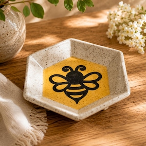 May include: A hexagonal ceramic dish with a speckled off-white finish and a central yellow honeycomb-shaped panel featuring a black bee illustration. The dish has raised edges and a smooth, glazed surface, designed as a small trinket tray.