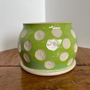 Puede incluir: Maceta de cerámica hecha a mano con un vibrante esmalte verde y grandes lunares blancos texturizados. La maceta tiene forma redondeada y se asienta sobre una pequeña base blanquecina. Ideal para plantas pequeñas o suculentas.