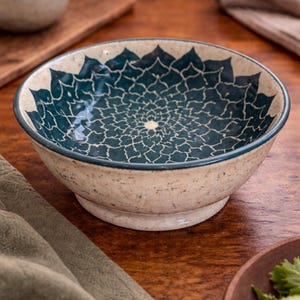 May include: Ceramic bowl with a teal and white floral pattern. The bowl has a beige exterior and a dark teal interior design. The bowl is on a wooden surface, with a green cloth and a small vase in the background.