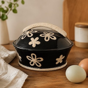 Puede incluir: Una mantequera de cerámica negra con un asa en la tapa y una base de color crema moteado. El exterior del recipiente y la tapa están decorados con motivos florales pintados a mano en color crema, cada uno con cinco pétalos dispersos.