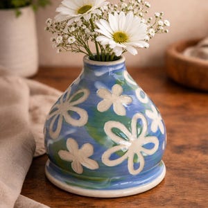 Könnte beinhalten: Eine blaue Keramikvase mit weißen Gänseblümchen und Schleierkrautblüten. Die Vase hat eine bauchige Form und ist mit weißen Blumenmustern verziert. Die Vase steht auf einer Holzoberfläche.