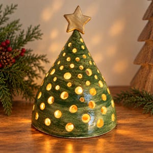 Puede incluir: Una decoración de árbol de Navidad cónica de cerámica con un esmalte verde moteado y numerosos recortes circulares que emiten una luz cálida. La pieza está rematada con una estrella de color dorado y tiene un acabado liso y brillante.