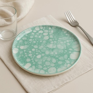 Op de afbeelding: Een ronde, turquoise keramische plaat met een wit bellenpatroon. Een helder glas en een zilveren vork zijn ook zichtbaar. De plaat ligt op een beige linnen servet.