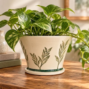 Può includere: Un vaso in ceramica color crema con bordo svasato e un anello di base verde scuro. L'esterno presenta motivi a rametti di foglie verdi dipinti a mano disposti verticalmente. Una rigogliosa pianta di pothos con foglie verdi e bianche.