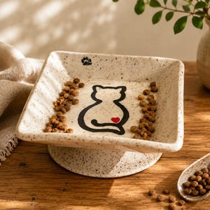 May include: A square, speckled ceramic pet bowl on a pedestal base. The interior features a black line drawing of a cat silhouette with a small red heart on its back and a tiny black paw print in the corner. The bowl contains brown dry pet food.
