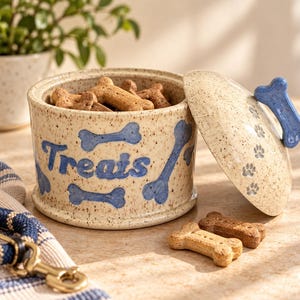 May include: A speckled ceramic treat jar with a removable lid, featuring the word "Treats" in blue script and blue bone illustrations. The lid has a blue bone-shaped handle and small paw print details. Two bone-shaped dog treats sit in front.