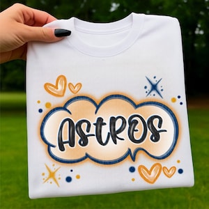 Puede incluir: Sudadera blanca de cuello redondo con la palabra "ASTROS" en un diseño en forma de nube. El diseño presenta colores naranja, azul y negro, con gráficos adicionales de corazones y estrellas. Una prenda casual y deportiva.
