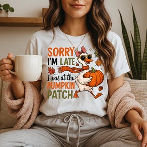 Könnte beinhalten: Weißes T-Shirt mit dem Text "SORRY I'M LATE I was at the PUMPKIN PATCH". Das Shirt zeigt eine Cartoon-Gans mit Sonnenbrille, Schal und Kürbis. Auf dem T-Shirt sind auch Herbstblätter aufgedruckt.