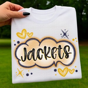 May include: White sweatshirt with the word "Jackets" in a black, stylized font inside a yellow and blue cloud-shaped design. The design includes yellow hearts and starbursts. The sweatshirt is held by a hand.