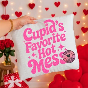 May include: White square pillow with the text "Cupid's Favorite Hot Mess" in bright pink. The pillow also has a small circular logo. The background includes red heart decorations.